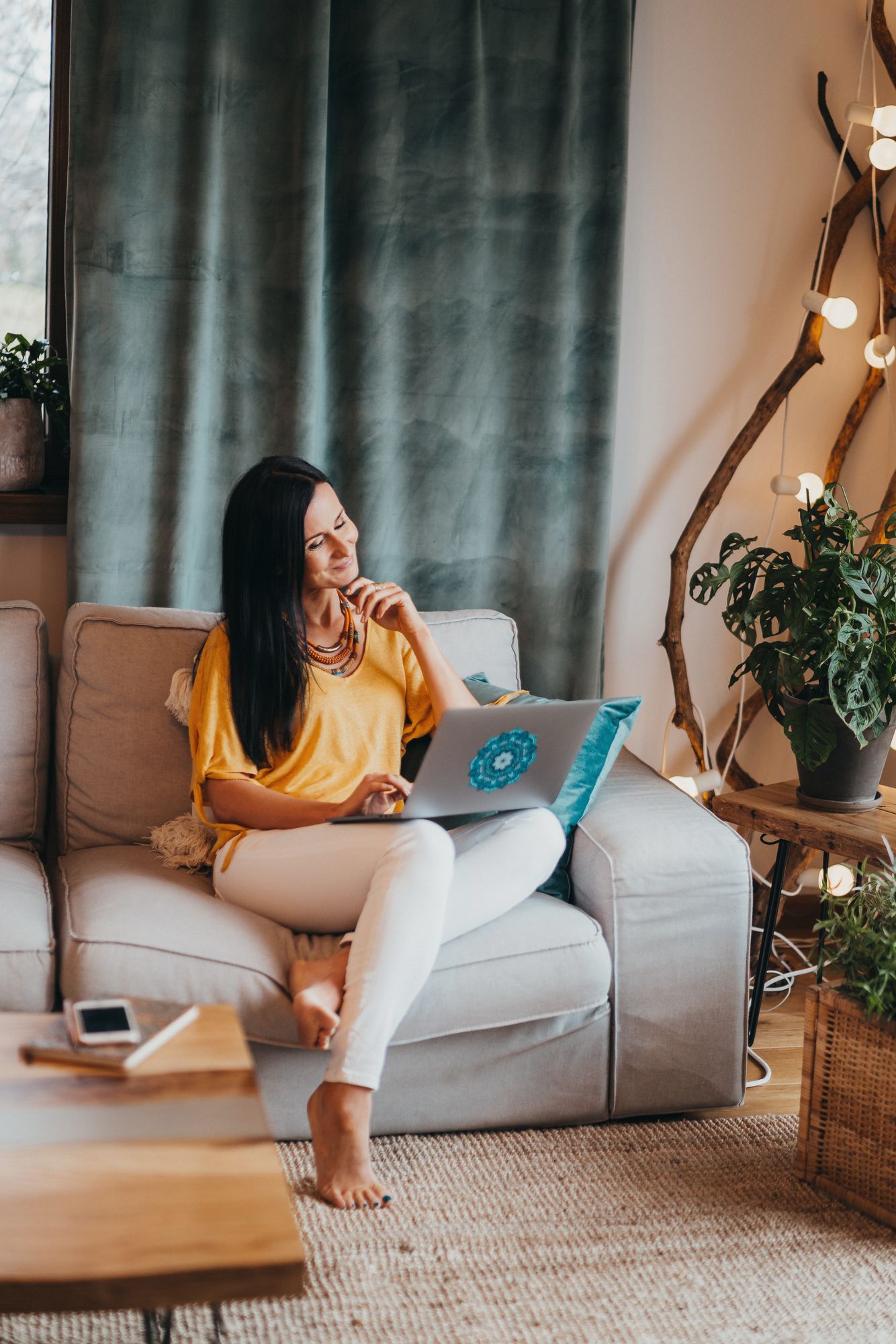 A female business coach sitting on a couch in a cozy living room, smiling and working on her laptop.